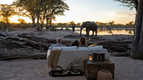 Tourists sitting at sundown watching an elephant drinking at the waterhole in front of a safari lodge