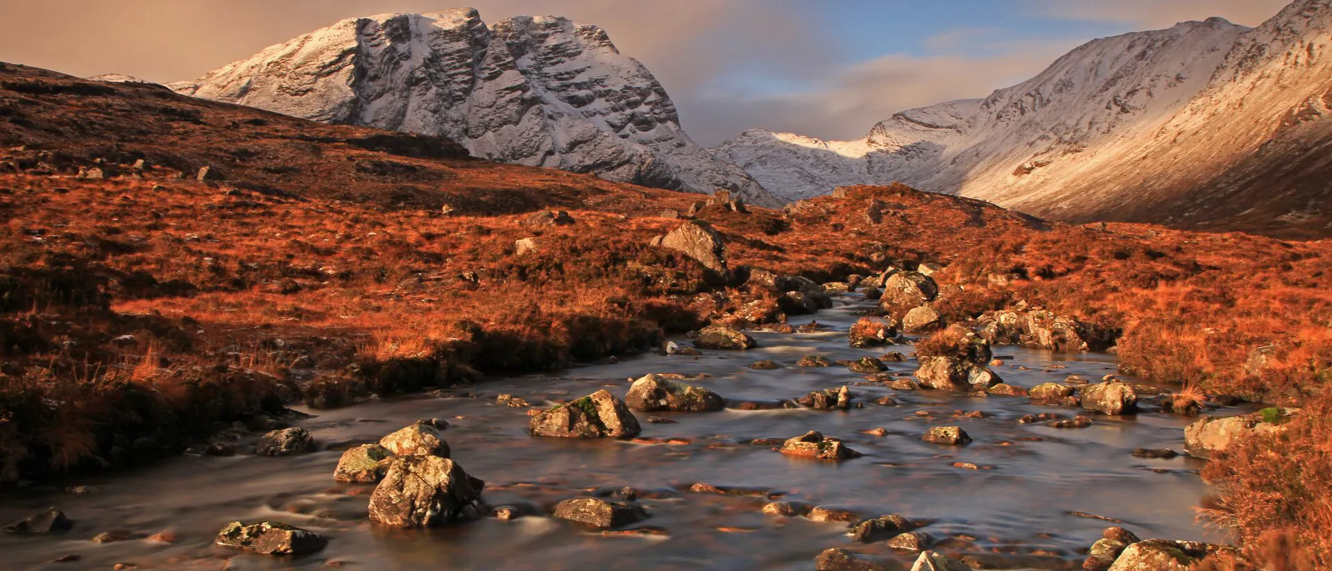 A winding river traverses a green valley, bordered by towering mountains under a clear blue sky