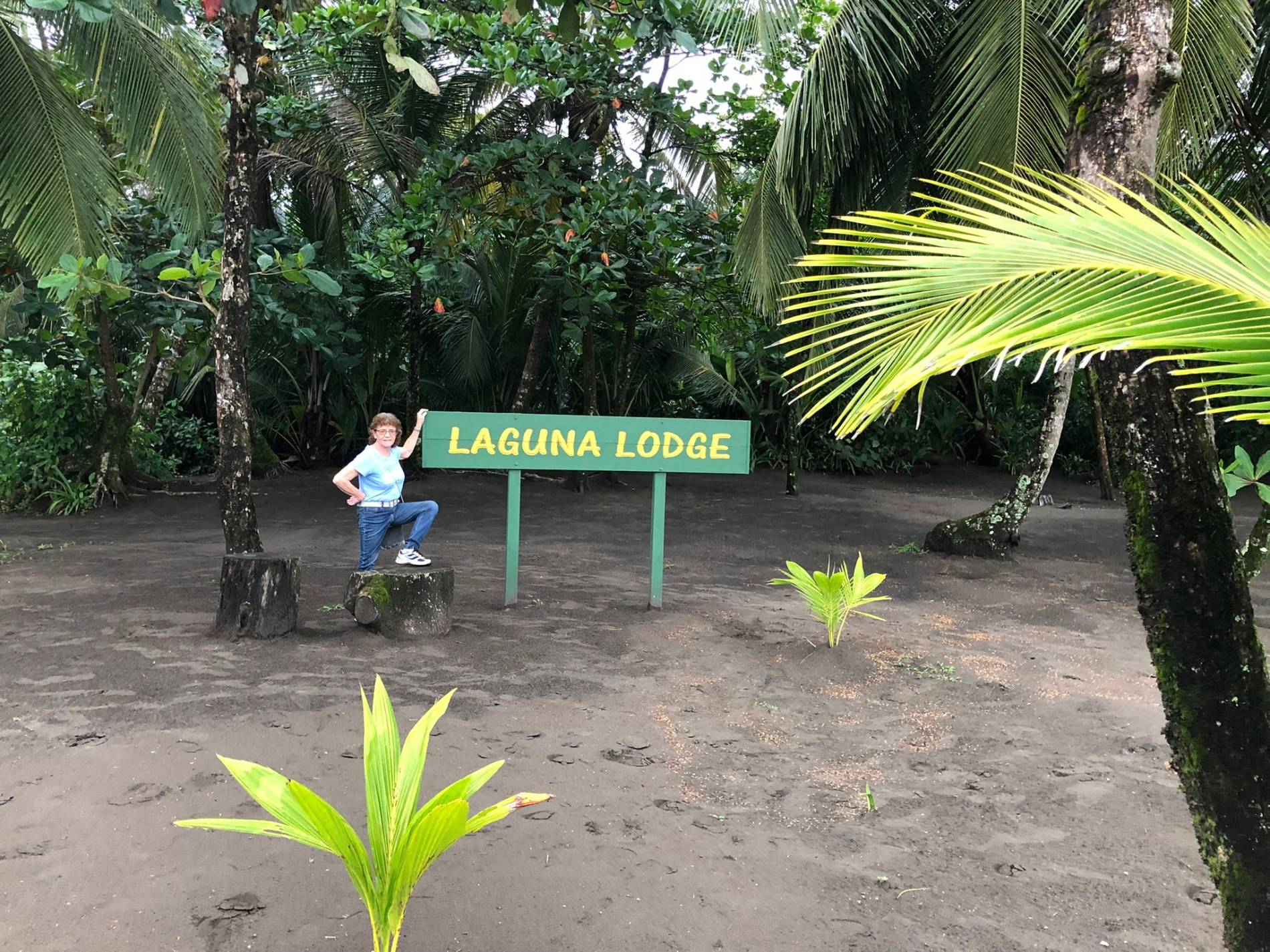 Anne Eyles standing next to Laguna Lodge Sign surrounded with troipical trees and bushes