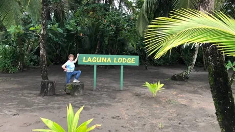 Anne Eyles standing next to Laguna Lodge Sign surrounded with troipical trees and bushes