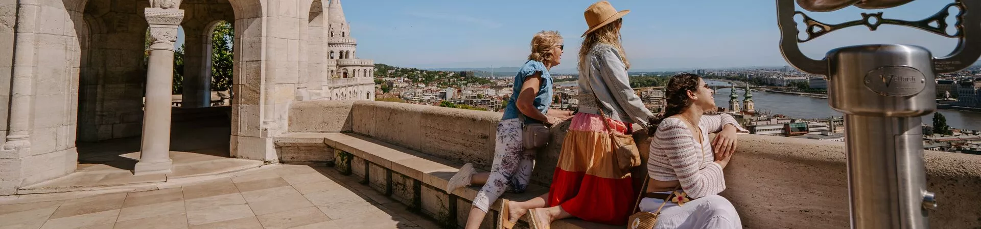 Three women look out over Budapest from an elevated stone balcony.