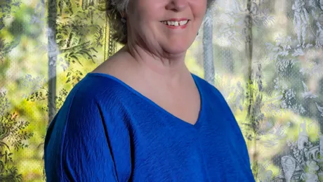 Marsha Colson Wearing a Blue Blouse Standing Next To Window
