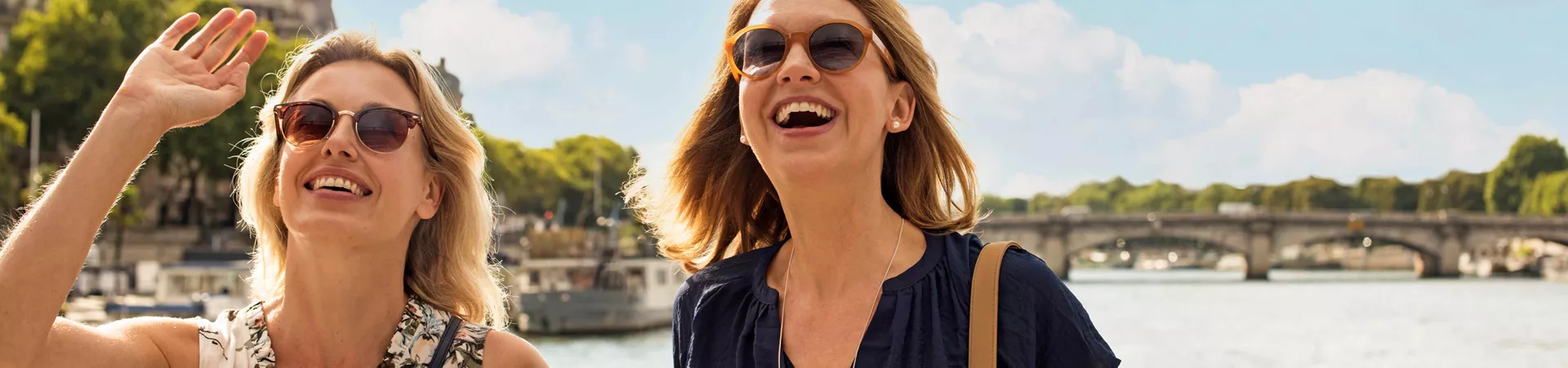 Two ladies in sunglasses smiling, stood in front of a river on a sunny day
