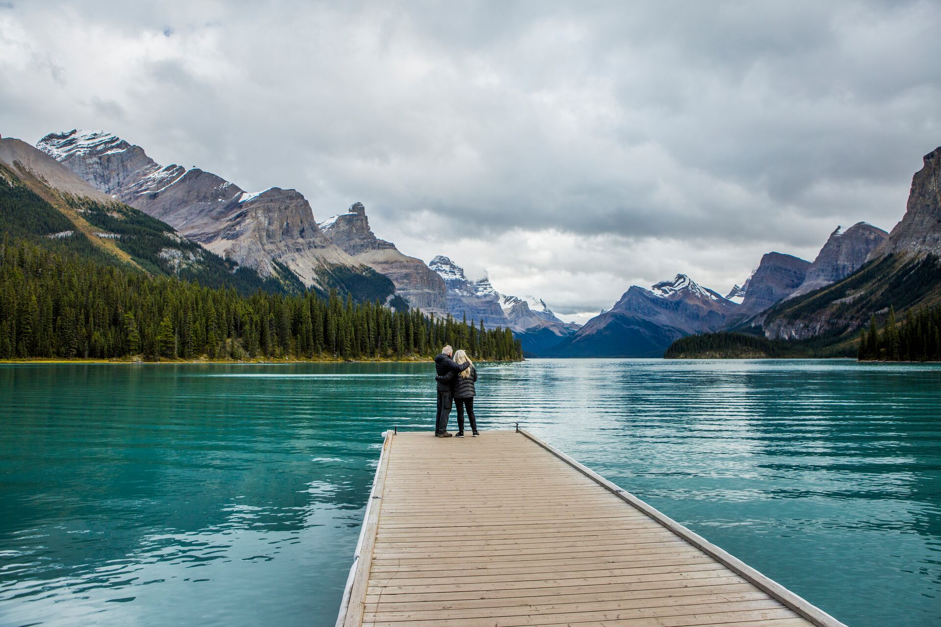 Couple Standing At The End Of Dock On Mountain Lake