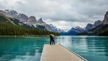 Couple Standing At The End Of Dock On Mountain Lake