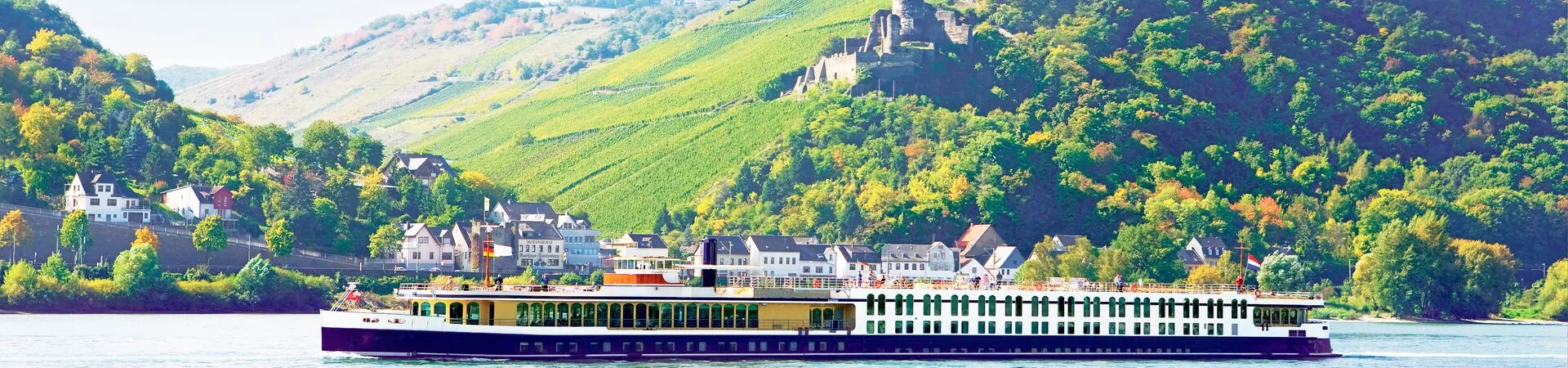 The river cruise Trafalgar Verity travels down the Rhine River with buildings along the shoreline and a castle above.