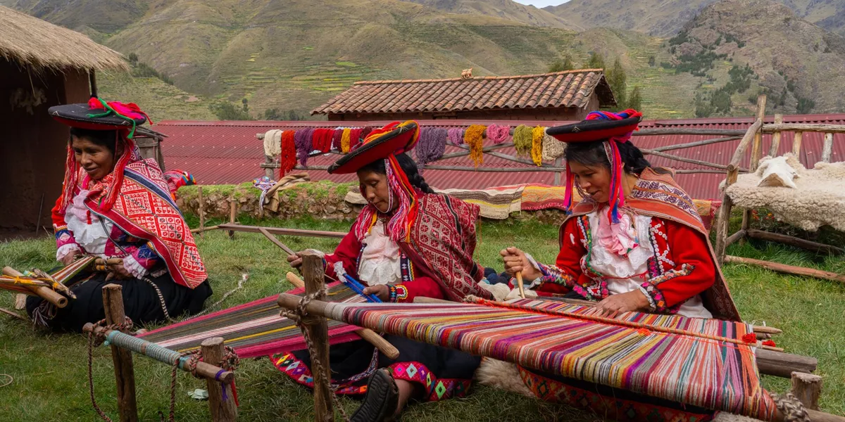 Three local female weavers in colourful traditional local dress including festooned hats, weaving colourful alpaca wool on the ground