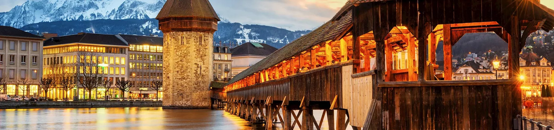 A bridge in Lucerne lit with warm light. A backdrop of snowy mountains.