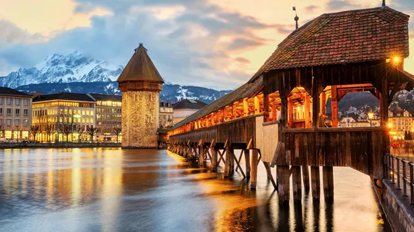 A bridge in Lucerne lit with warm light. A backdrop of snowy mountains.