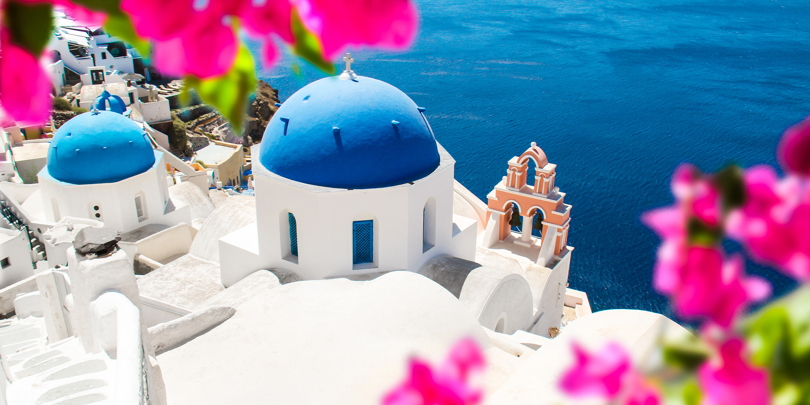 A white building with blue domed roof framed by pink flowers in Santorini, Greece