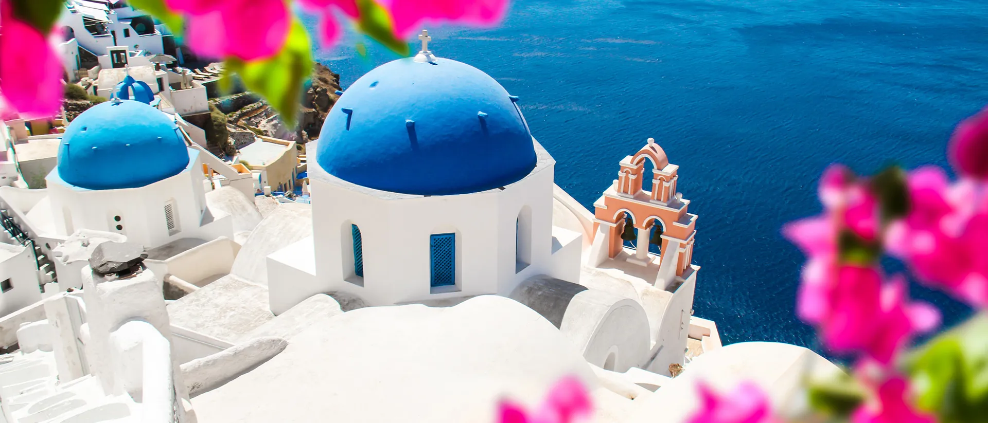 A white building with blue domed roof framed by pink flowers in Santorini, Greece