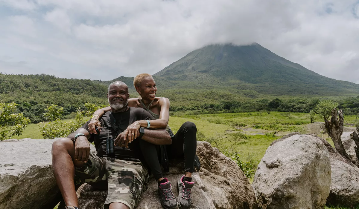 A man and woman sitting on a rock with Arenal Volcano in the background