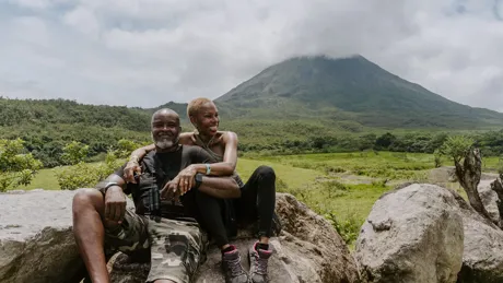 A man and woman sitting on a rock with Arenal Volcano in the background