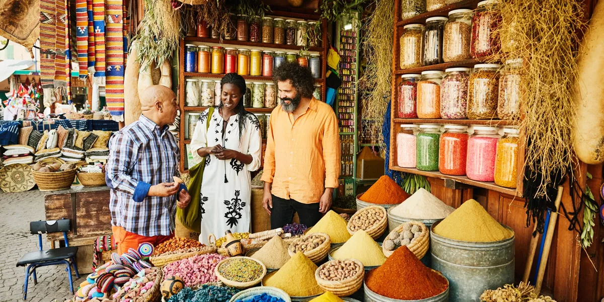 Wide Shot Couple Talking To Spice Shop Owner In The Souks Of Marrakech, North Africa
