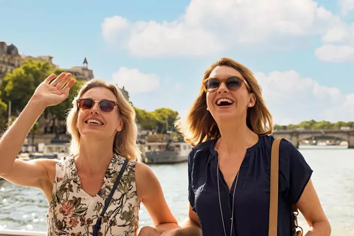 Two ladies in sunglasses smiling, stood in front of a river on a sunny day
