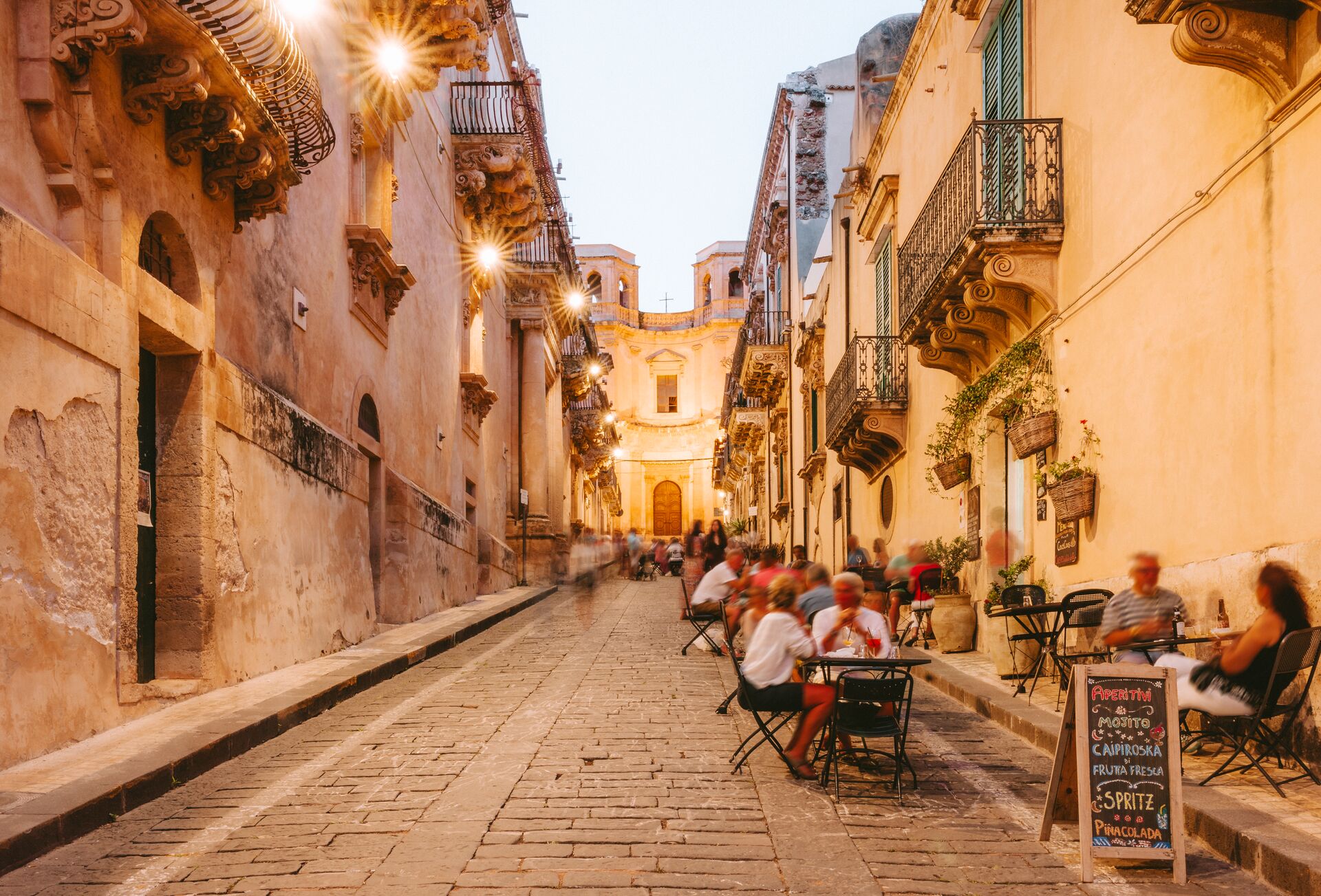 People dining outside restaurants and bars at dusk in Noto, Italy