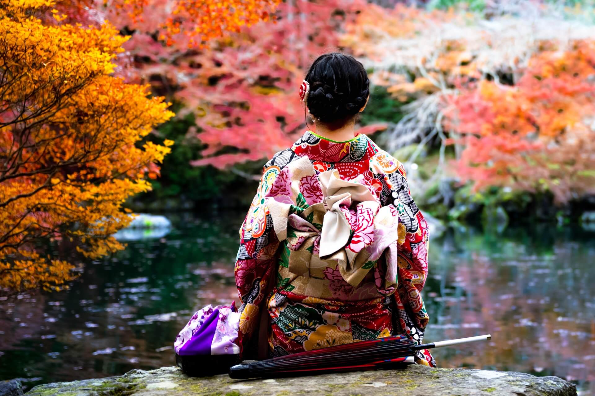 Woman in a colorful kimono sits by a pond surrounded by vibrant autumn leaves