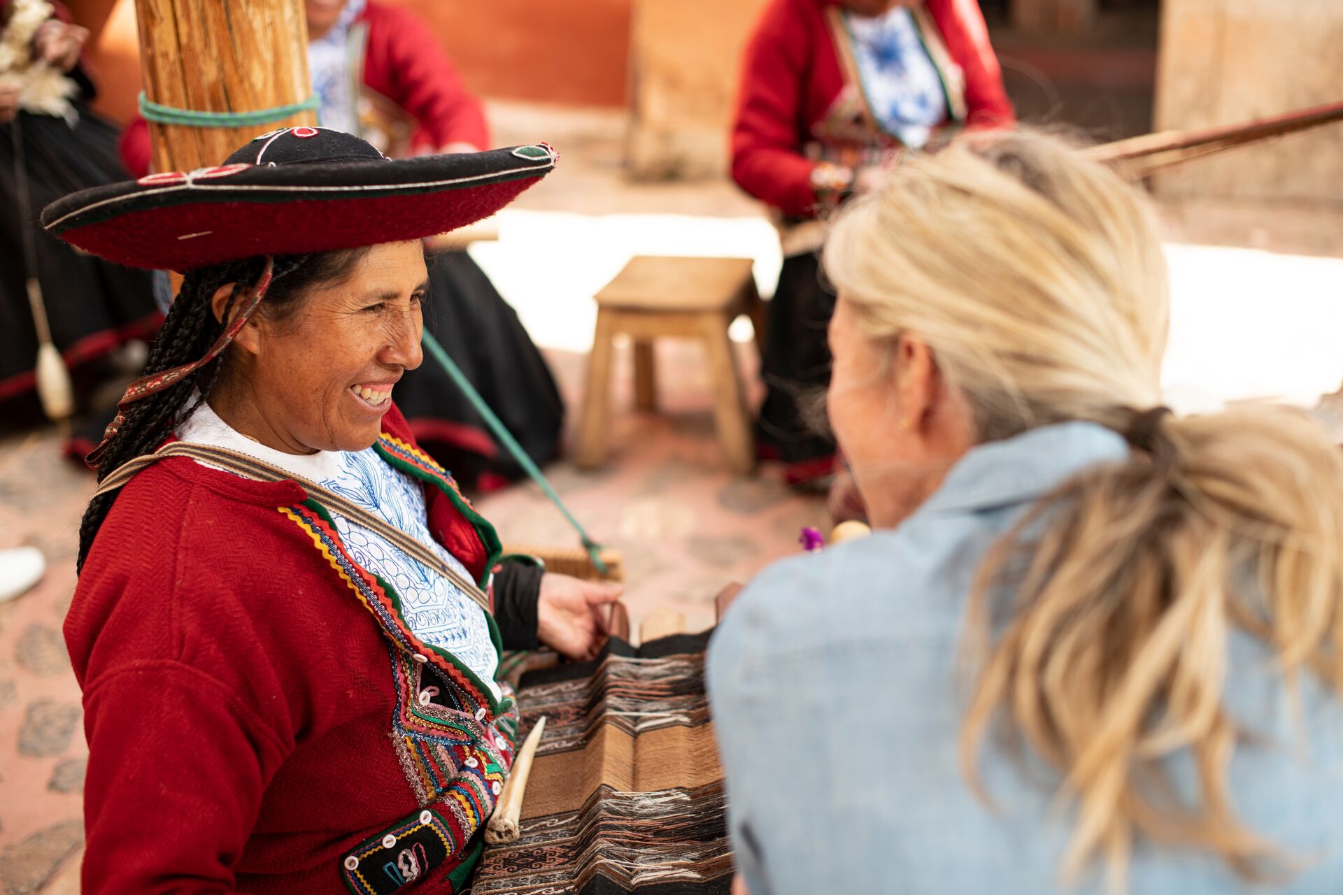 Woman in traditional Peruvian outfit knitting and smiling while talking with blonde woman