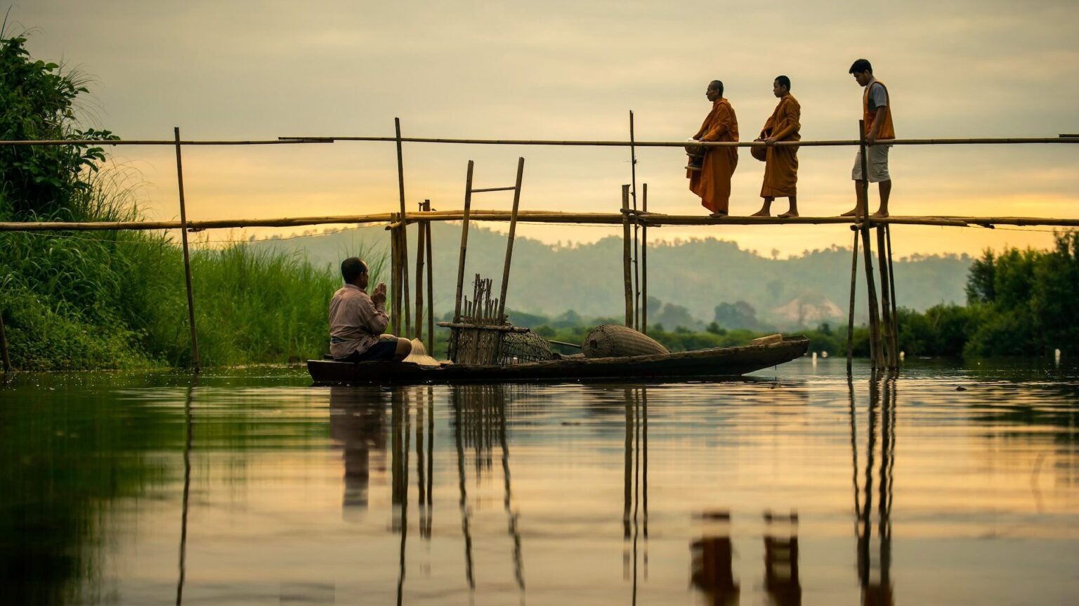 Two Buddhist monks and a man walk on a bamboo bridge over a calm river in rural Thailand at sunrise, while another man sits in a wooden boat below