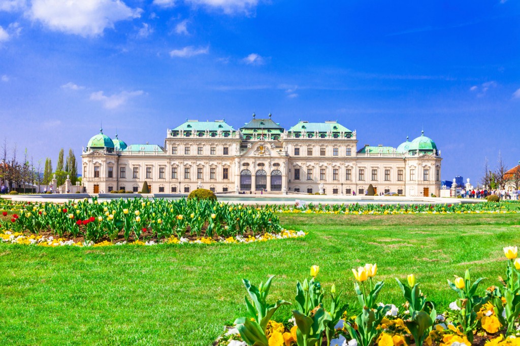 Belvedere Palace in Vienna during springtime. Yellow tulip flowers bloom on its grounds and the sky is a clear blue with only a few wisps of cloud.