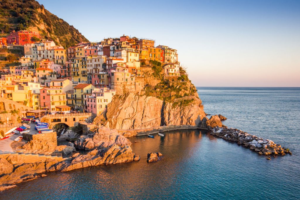 A seaside town in Italy in September basks in golden hour light, the multi-colored houses bright and welcoming. The sea is flat and the sky has an orange to blue gradient.
