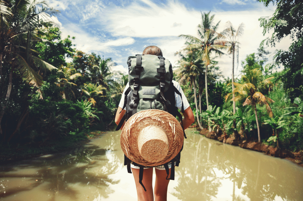 person with a large backpack and straw hat stood with their back to the camera in front of a creek/river. Trees line the sides of the water.