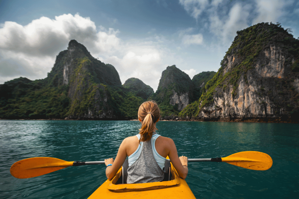 Woman kayaking in the sea, with large mountainous rock formations ahead of her along with a small beach.