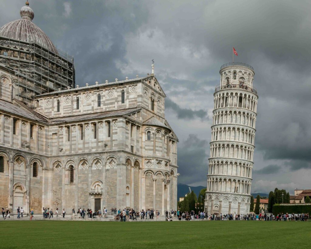 Dark cloudy sky, with the Leaning Tower of Pisa building, Italy, visible in the center-right. This is a major European landmark. To the left, is a grand cathedral. Grass in the forefront, with tourists stood outside the buildings in the background.