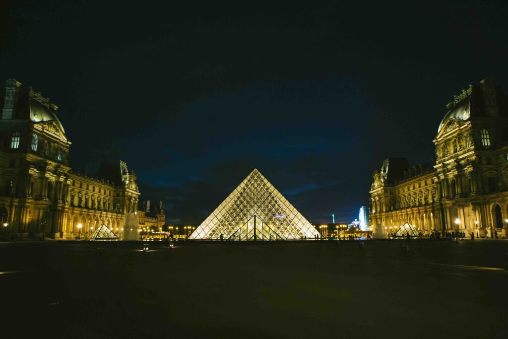 The Louvre Museum in Paris, France, illuminated at night, with its glass pyramid glowing warmly against the dark sky. European landmark.