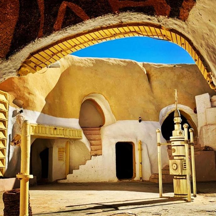 Looking through an archway we see the courtyard of this Star Wars filming location. A droid stands in the courtyard under a blue sky and doorways carved into the stone wall lead to different chambers