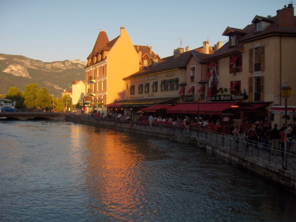 Canal/river on the left hand side side, with it lined by pastel coloured buildings on the right-hand side. Outside dining is visible outside of the buildings.