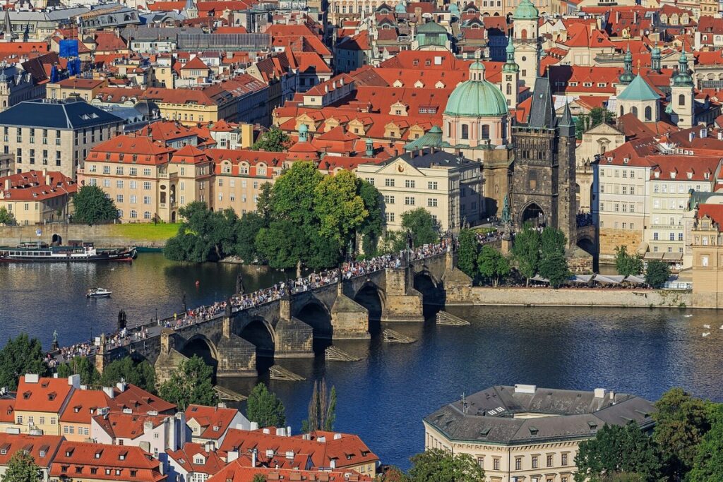 Birds-eye view of Charles Bridge in Prague, the bridge is full of people walking across it. River underneath and red roof buildings in the background. Sunny day.