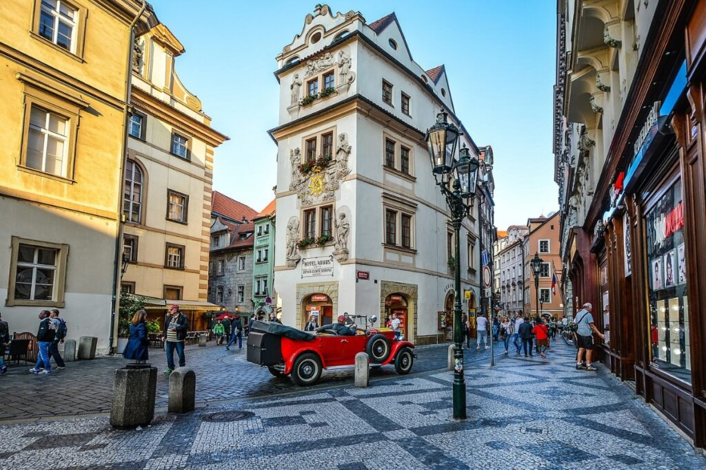 Cobblestone old-town square in Prague, with a red vintage car in the middle. Buildings surround the car, blue skies.