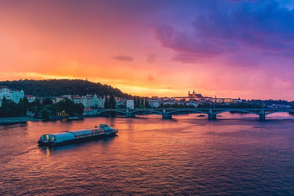 Sunset skies reflecting on the river in Prague, bridge in the background and a boat on the river.