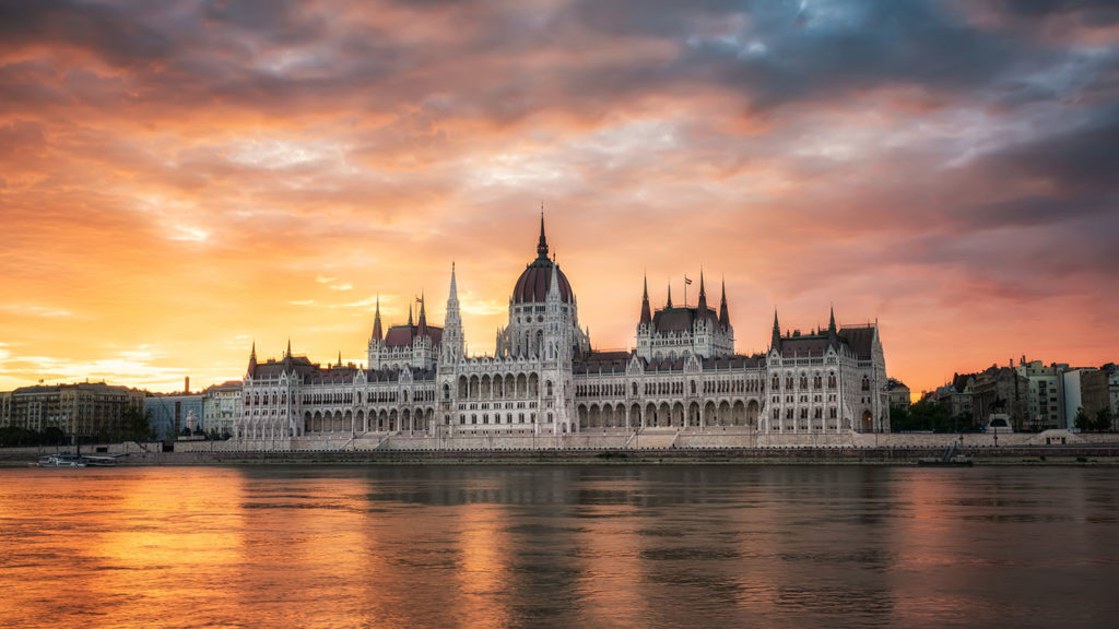 Domed and spired building on the bank of a river, with a sunset-hued sky behind