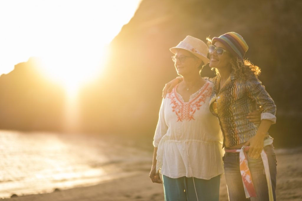 Mother and daughter caucasian people friends at the beach hugging and having fun smiling with golden sun in backlight and ocean in background - family and mixed diversity generations concept