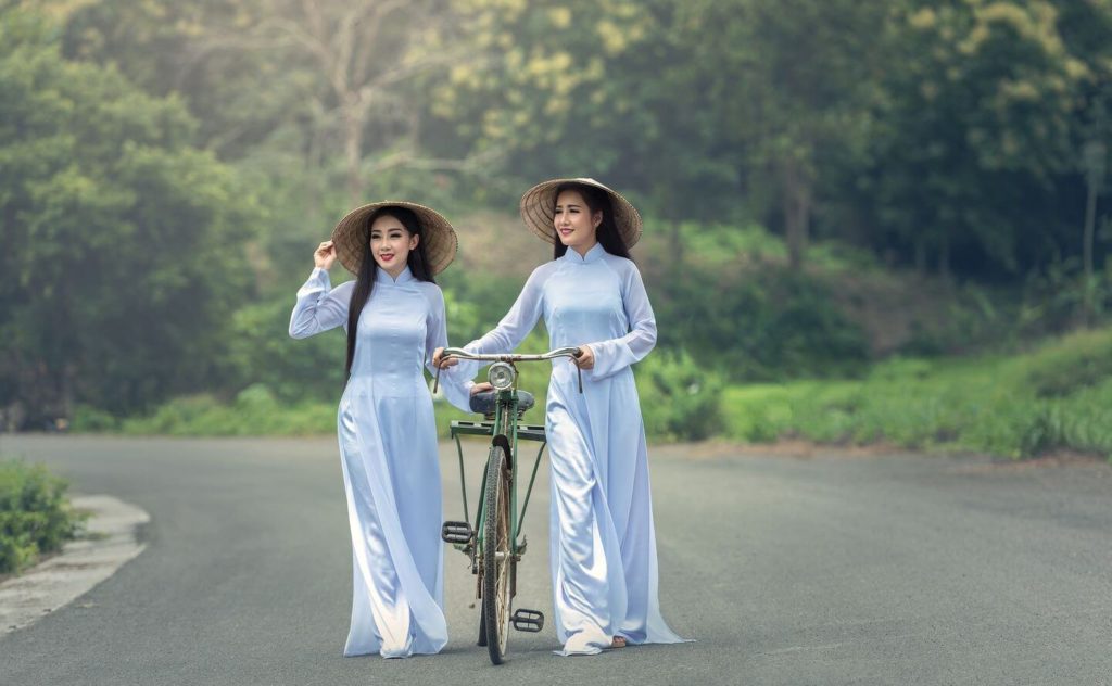 Two women in traditional Vietnamese dresses and conical hats standing with a bicycle on a road surrounded by greenery.