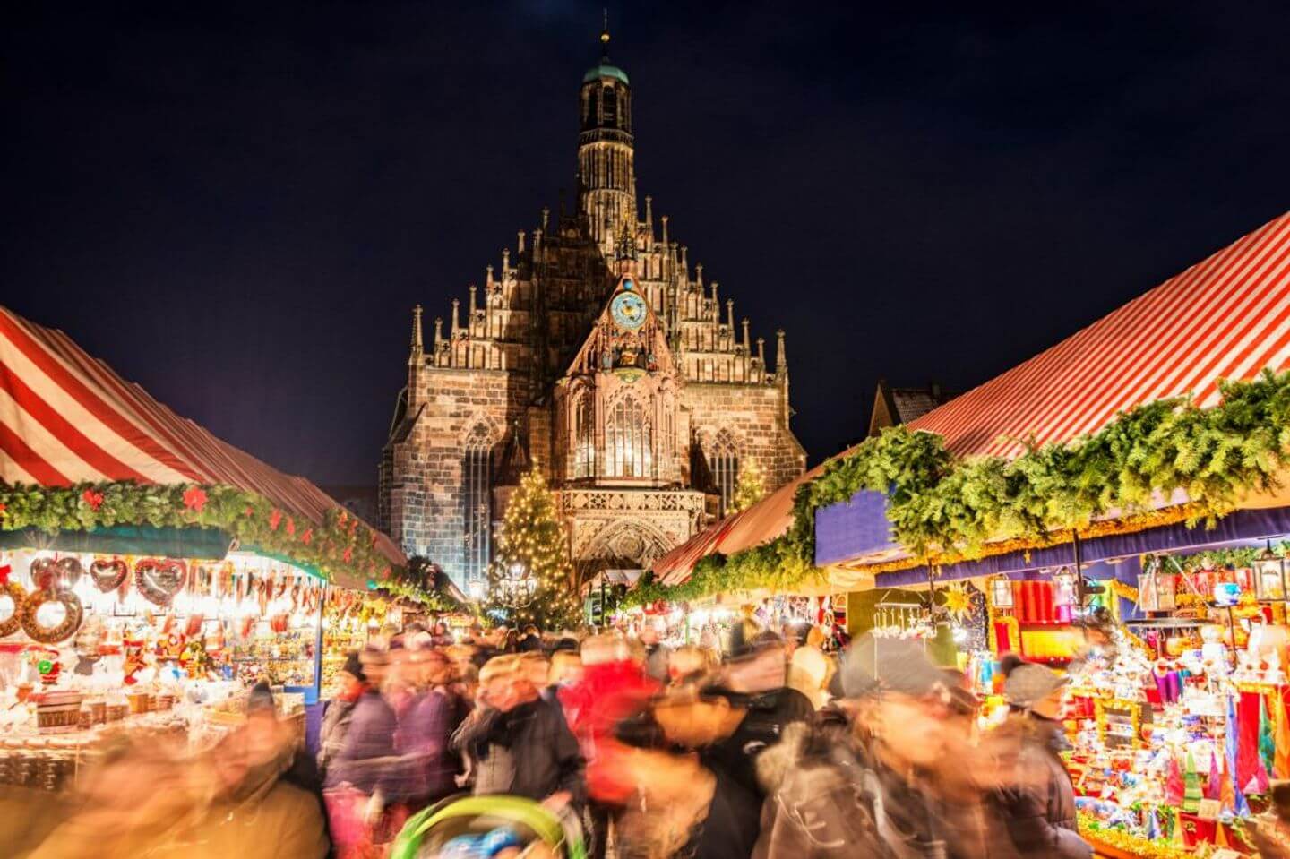 A bustling German Christmas market at night with blurred figures of people shopping, set against the illuminated backdrop of a gothic cathedral.