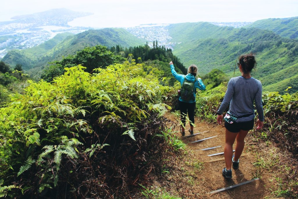 Two hikers on a forest trail in Hawaii, with one pointing towards a scenic overlook.