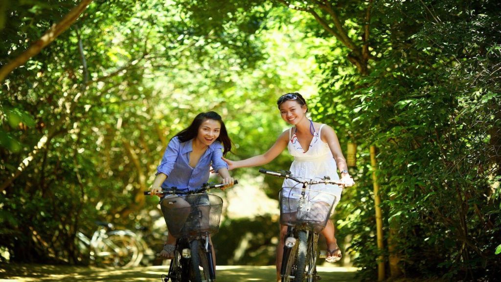 two women smiling and cycling down a leafy street