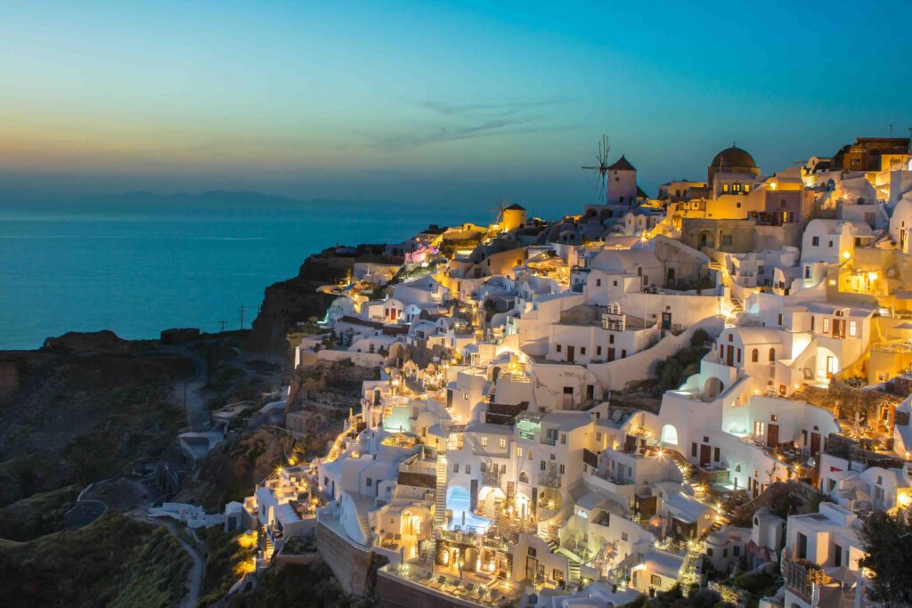 Oia in Greece, many buildings on the side of a cliff. White buildings, some with lights illuminating the houses/hotels. Can see the sea in the distance on the left.