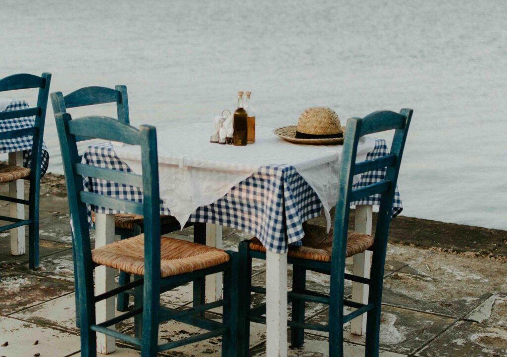 Small white table with blue and white checkered tablecloth on it. Three blue chairs are tucked under the table, with two oil dispensers on the top alongside a straw hat.