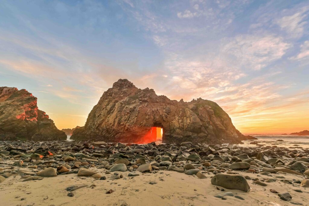 Pfeiffer beach, sandy beach with large rocks. There's a large rock formation coming out of the water, with the sun setting peaking through.