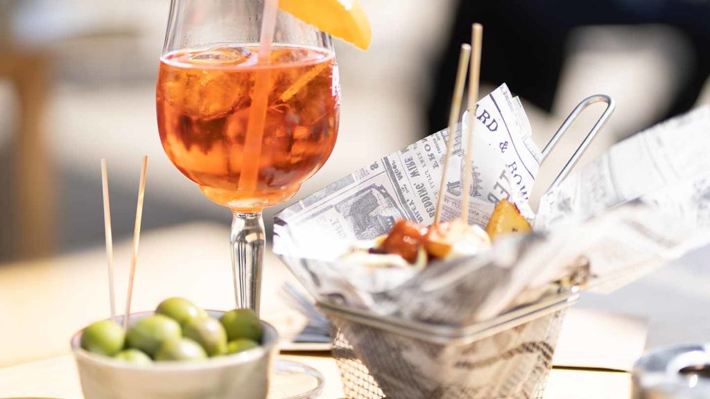 An aperol spritz accompanied by olives and snacks served in a newspaper-lined cone, presented on a sunny outdoor table.