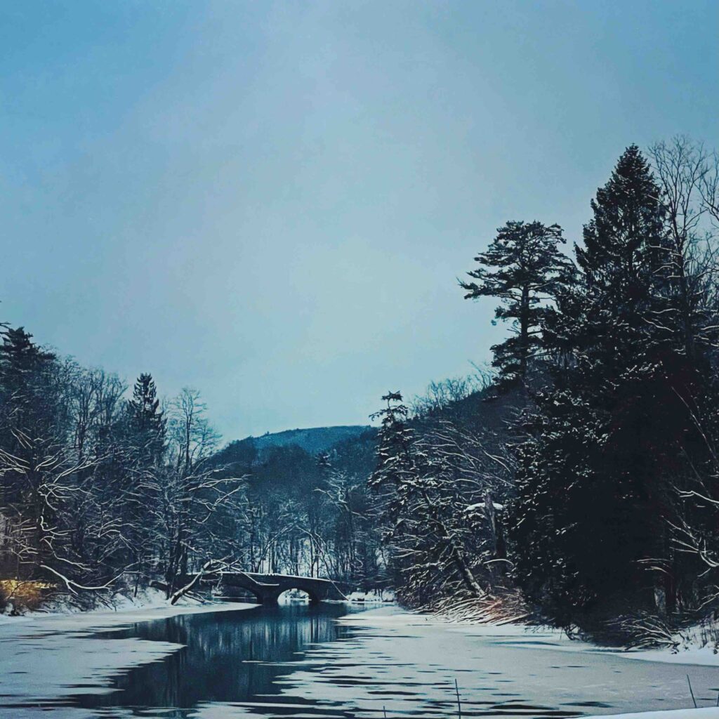Cooperstown New York, winter time, with trees on the outsides of the image and an icy lake running through the center. A stone bridge is in the background. family Christmas vacation ideas