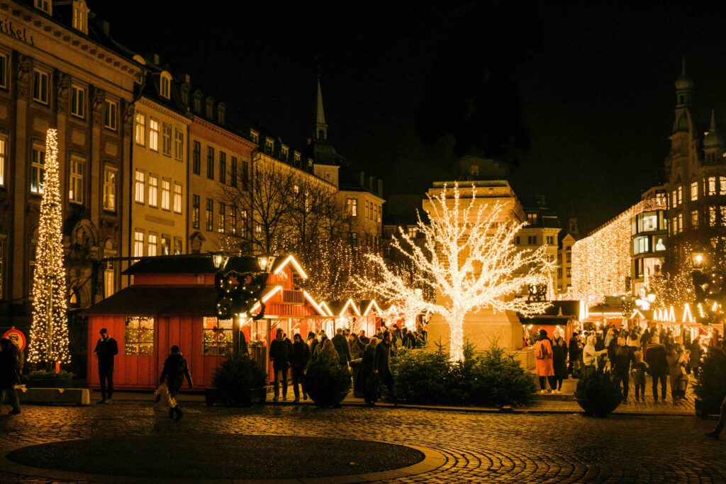 Christmas market in Copenhagen, buildings are lining the street with Christmas lights lighting up the trees dotted around and the small cabins.