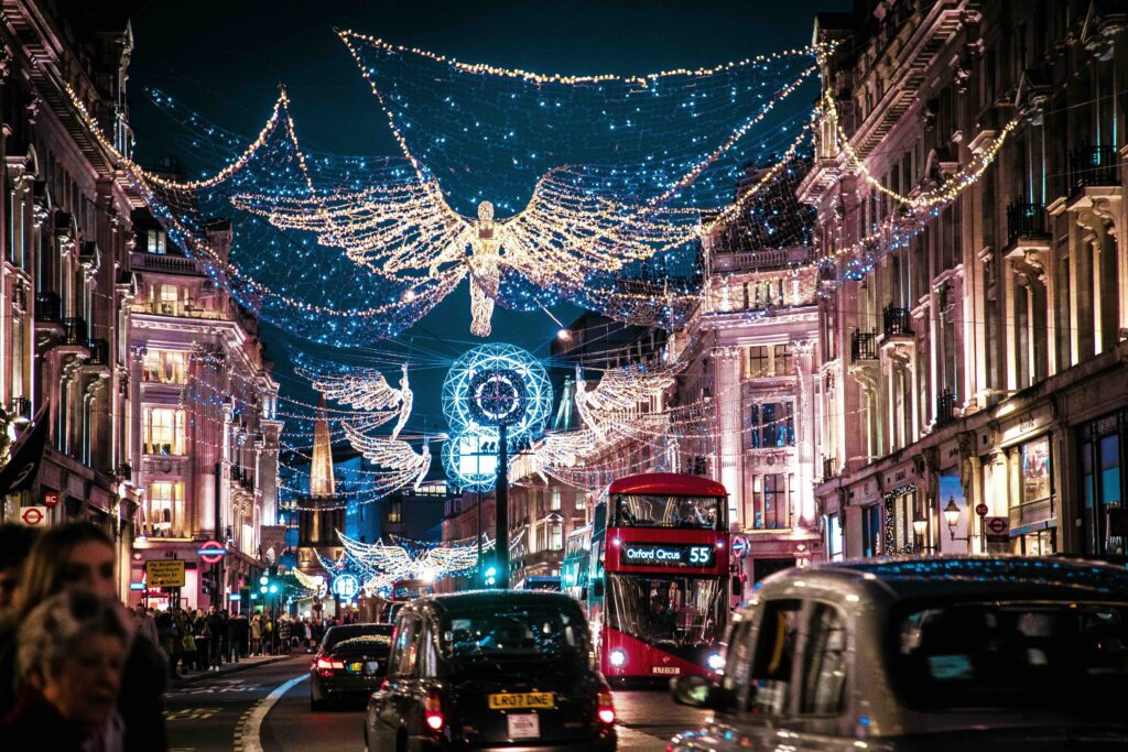 Oxford Street in London, family Christmas vacation ideas. Stores are lining the sides of the images, with traffic on the road in the center. Double-decker bus coming towards the camera, Christmas lights above the street.