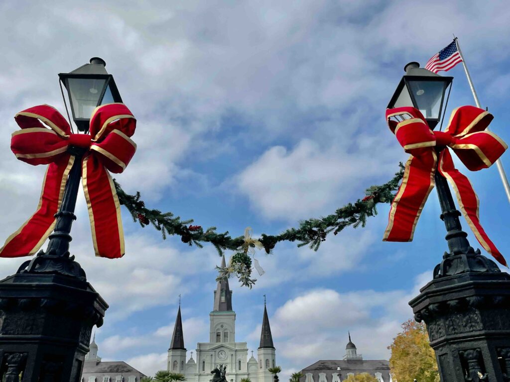 Close-up of two lamp posts with large red and gold Christmas bows tied on them and holly attached to them both. Can see buildings in the background