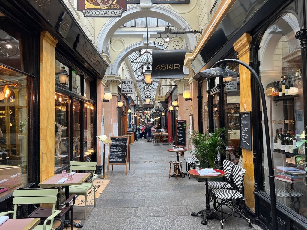 Inside Passage des Panoramas in Paris, stone floor, chairs sat outside cafes/restaurants, with the signs of shops attached to the buildings.