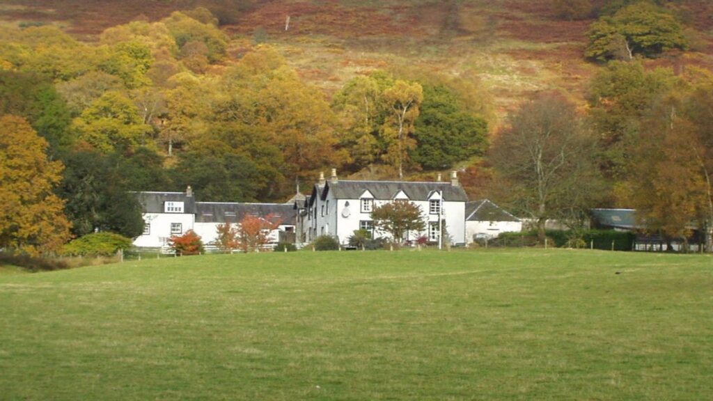 A rural landscape with a white Scottish farmhouse surrounded by autumn-colored trees and a grassy field in the foreground.